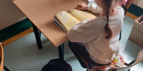 Powiększ grafikę: Kwadratowy stolik , przy którym siedzą dwie dziewczynki i czytają książki. Dziewczynka po prawej stronie ma warkocze. Czytana przez nią książka ma żółte kartki i białe obrazki. 