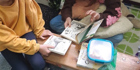 Powiększ grafikę: Dwie dziewczynki czytają książki. Dziewczynak po lewej stronie siedzi na ławeczce gimnastycznej. Ma żółtą bluzkę i niebieskie spodnie. Obok niej leży kolejna książka, na której stoi niebiesko-biała śniadaniówka. Druga dziewczynka siedzi po turecku, za ławeczką. Na kolanach trzyma książkę. Na lewym kolanie dziewczynki leży szara poduszka. Po prawej stronie leży czarny plecak. 