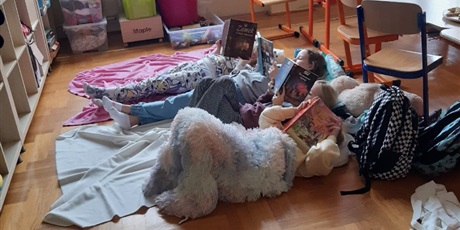 Powiększ grafikę: Sala lekcyjna. Na parkiecie leżą koce. Na kocach leży troje dzieci czytających książki. Głowy dzieci zwrócone są w prawą stronę. Na pierwszym planie dziewczynka otulona jasno noebieskim kocykiem. Zasłania twarz książką z różową okładką. Obok niej dwie dziewczynki w niebieskich spodniach.  Leżą na niebieskiej poduszcze. Na końcu dziewczyn ka w kolorowej piżamce. Leży na kolorowym kocu. Za głowami dziewczynek widać krzesełka szkolne i fragment ławki. 