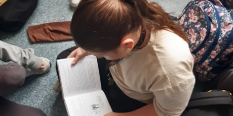 Powiększ grafikę: Dziewczynka w jasnej koszulce i ciemnych włosach związanych w kitek, siedzi na podłodze i czyta książkę z pochyloną głową. Obok dziewczynki stoi kolorowy plecak. 
